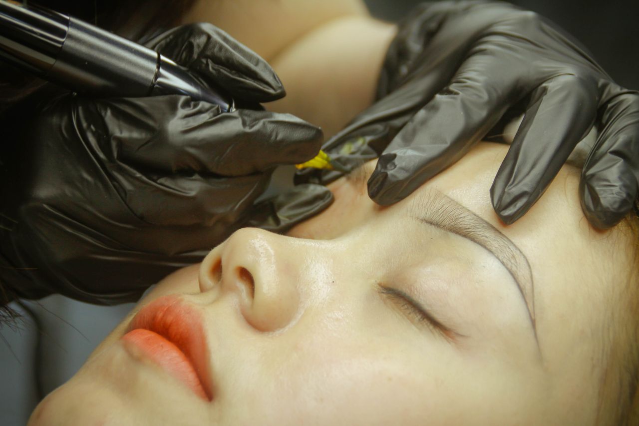 services-03 A detailed view of eyebrow microblading procedure with gloved hands and closed eyes.