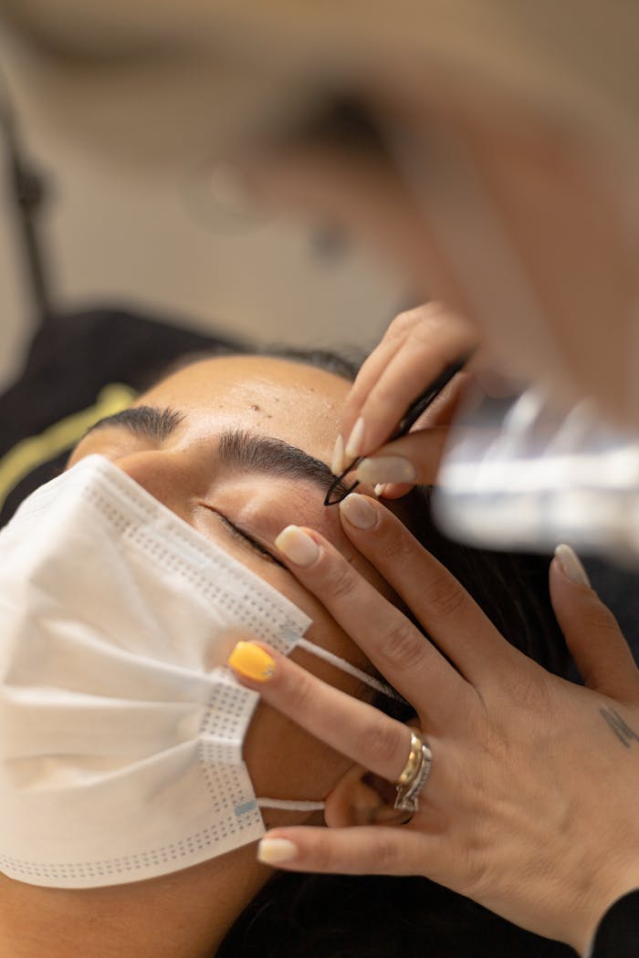about-bg A woman with closed eyes wearing a mask receives a beauty treatment for her eyebrows.