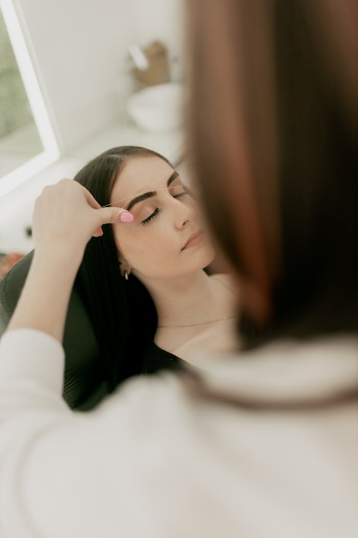 services-04 A beautician shaping a young woman's eyebrows in a serene salon setting.